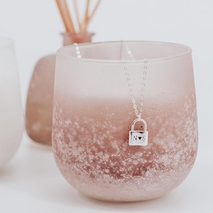 Puede incluir: Un collar de plata con un colgante de candado. El candado tiene las iniciales "N" y "S" grabadas, con un pequeño símbolo de corazón entre las letras.