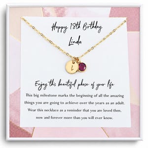 May include: A gold-tone chain necklace featuring a small circular pendant engraved with the letter L and a round dark pink gemstone charm. The necklace is displayed on a white card with the text Happy 18th Birthday Linda and a printed message.