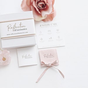 May include: A pink jewelry box with the text "Reflection of memories" and a website address. The box is open and a gold necklace with a pink crystal pendant is visible. There is a pink velvet pouch with the same text on it, and a card with the same text. The image also includes a jewelry care instruction card with icons.