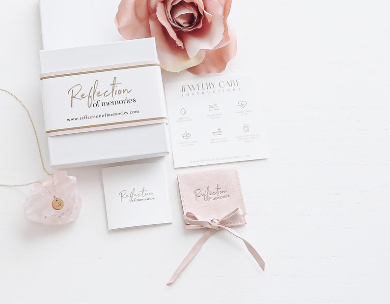 May include: A pink jewelry box with the text "Reflection of memories" and a website address. The box is open and a gold necklace with a pink crystal pendant is visible. There is a pink velvet pouch with the same text on it, and a card with the same text. The image also includes a jewelry care instruction card with icons.