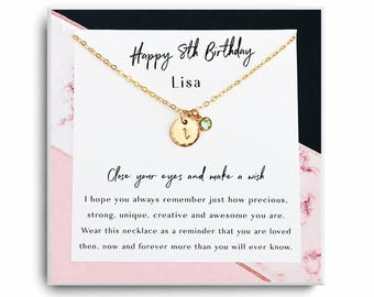 May include: A gold chain necklace featuring a small, round, hammered gold charm with the letter L and a tiny green crystal charm. The necklace is displayed on a white card with the text Happy 8th Birthday Lisa and a printed message about making a wish.