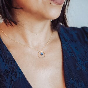 Puede incluir: Un collar de oro con una cadena delicada y un peque&ntilde;o colgante de piedra azul que cuelga de un c&iacute;rculo.