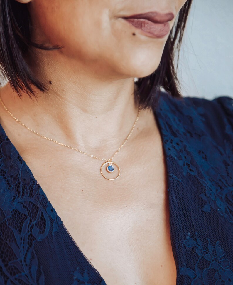 Puede incluir: Un collar de oro con una cadena delicada y un peque&ntilde;o colgante de piedra azul que cuelga de un c&iacute;rculo.