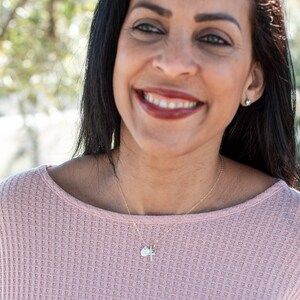 May include: A woman wearing a pink waffle knit sweater with a gold chain necklace with multiple charms. The charms include a small round charm, a cross charm, and a small green stone charm.