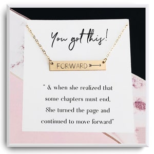 Op de afbeelding: Gouden ketting met een hanger in de vorm van een staaf waarop "FORWARD" staat met een pijl die naar rechts wijst. De ketting zit in een witte doos met een roze-wit marmerpatroon. De doos bevat ook een citaat dat luidt: "& toen ze besefte dat sommige hoofdstukken moesten eindigen, sloeg ze de pagina om en ging ze verder."