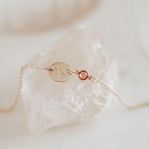 Peut inclure: Un bracelet d&eacute;licat en cha&icirc;ne dor&eacute;e avec un petit charme rond grav&eacute; de la lettre "K" et une petite pierre pr&eacute;cieuse rouge.