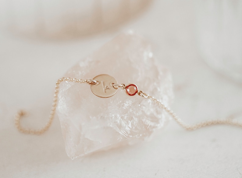 Peut inclure: Un bracelet d&eacute;licat en cha&icirc;ne dor&eacute;e avec un petit charme rond grav&eacute; de la lettre "K" et une petite pierre pr&eacute;cieuse rouge.