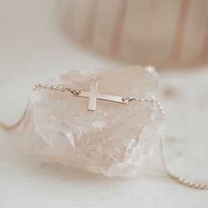 May include: A delicate gold chain necklace with a simple, flat cross pendant. The necklace is displayed on a pink crystal.