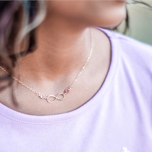 May include: A gold infinity necklace with two small gemstones, one pink and one clear, on a person wearing a light purple shirt.