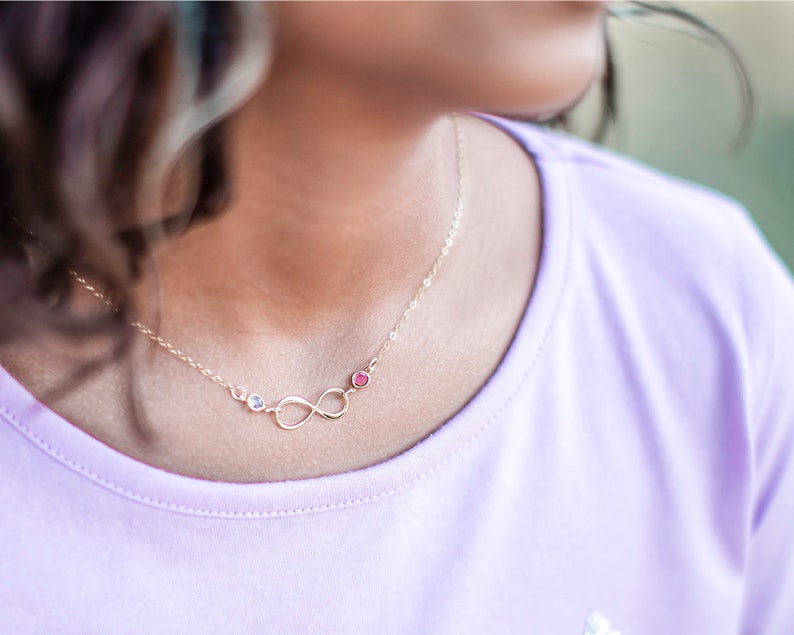 May include: A gold infinity necklace with two small gemstones, one pink and one clear, on a person wearing a light purple shirt.
