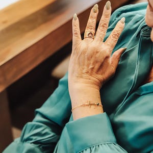 Puede incluir: Un anillo de oro con un dise&ntilde;o en forma de coraz&oacute;n y una pulsera de oro con un dije de cruz. La pulsera tambi&eacute;n presenta peque&ntilde;as cuentas rosas y verdes. La joyer&iacute;a se usa en una mano con u&ntilde;as largas y pintadas.