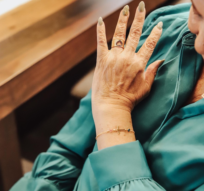 Puede incluir: Un anillo de oro con un dise&ntilde;o en forma de coraz&oacute;n y una pulsera de oro con un dije de cruz. La pulsera tambi&eacute;n presenta peque&ntilde;as cuentas rosas y verdes. La joyer&iacute;a se usa en una mano con u&ntilde;as largas y pintadas.