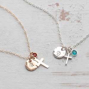 May include: Two delicate necklaces with silver and gold chains. Each necklace features a small cross charm, a hammered metal disc with an initial, and a gemstone. The necklace on the left has a red gemstone, while the necklace on the right has a turquoise gemstone.