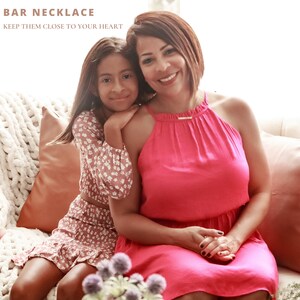 May include: A mother and daughter are sitting on a couch. The mother is wearing a pink halter dress and a gold bar necklace. The daughter is wearing a floral print dress. The text "BAR NECKLACE KEEP THEM CLOSE TO YOUR HEART" is at the top of the image.