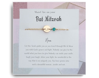 May include: A gold chain bracelet with a small round charm engraved with the letter "N" and a teal blue gemstone. The bracelet is displayed on a white card with the text "Mazel Tov on your Bat Mitzvah Noa Let the Torah guide you as you travel through life & bless you with God's peace and light. Nobody can give to the world what you have to give. Nobody can smile your smile, laugh your laugh, shine your light, be wonderful in the way that is so uniquely you. You have grown into such a beautiful woman...inside and out."