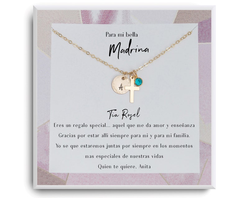 May include: A gold necklace with a cross charm, a small initial charm, and a turquoise gemstone. The necklace is in a white box with a pink and white patterned background. The box has a message written in Spanish that says "Para mi bella Madrina" and "Eres un regalo special... aquel que me da amor y ense&ntilde;anza Gracias por estar alli siempre para mi y para mi familia. Yo se que estaremos juntas por siempre en los momentos mas especiales de nuestras vidas Quien te quiere. Anita"