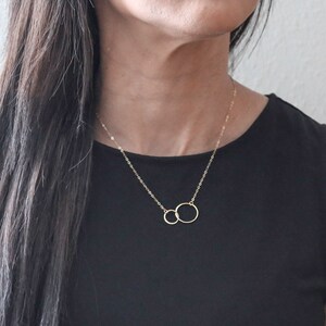 May include: A gold necklace with two interlocking circles. The necklace is on a person wearing a black top.
