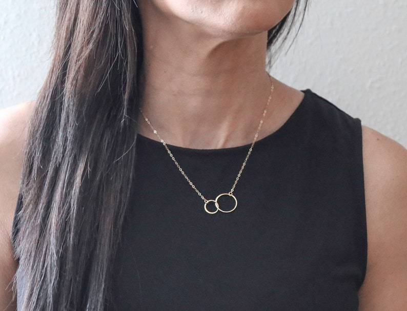 May include: A gold necklace with two interlocking circles. The necklace is on a person wearing a black top.