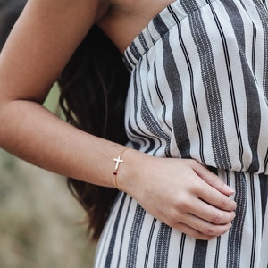 Puede incluir: Una pulsera dorada con un colgante de cruz y una peque&ntilde;a gema roja. La pulsera se lleva en la mu&ntilde;eca, con un mono a rayas blancas y negras de fondo.