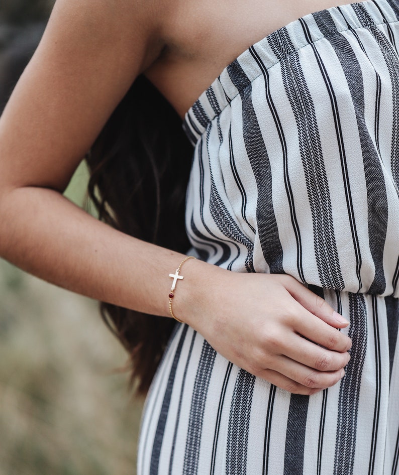 Puede incluir: Una pulsera dorada con un colgante de cruz y una peque&ntilde;a gema roja. La pulsera se lleva en la mu&ntilde;eca, con un mono a rayas blancas y negras de fondo.