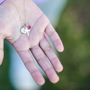 May include: A gold chain necklace with a small cross charm and a round charm with a letter on it. The charm has a small pink gemstone on it.