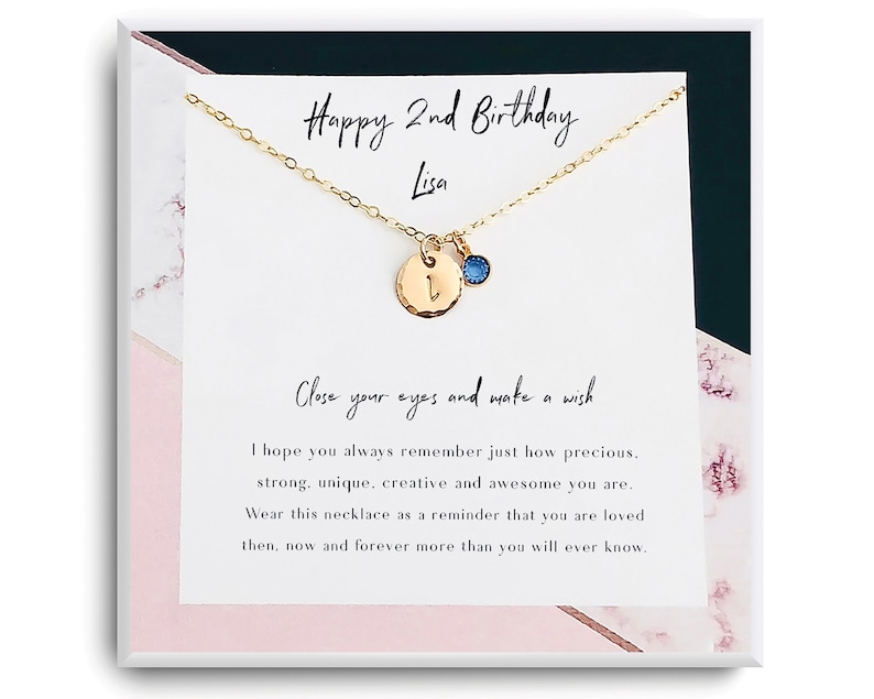 May include: A gold necklace with a small charm featuring the letter "L" and a blue gemstone. The necklace is presented in a white box with a pink and white marble design. The box includes a message that reads "Happy 2nd Birthday Lisa. Close your eyes and make a wish. I hope you always remember just how precious, strong, unique, creative and awesome you are. Wear this necklace as a reminder that you are loved then, now and forever more than you will ever know."