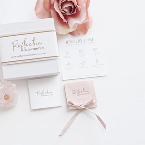 May include: A pink jewelry box with the text "Reflection of memories" and a website address. The box is on a white surface with a pink rose, a small pink pouch with a ribbon, and a necklace with a pink crystal pendant.