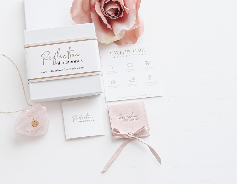 May include: A pink jewelry box with the text "Reflection of memories" and a website address. The box is on a white surface with a pink rose, a small pink pouch with a ribbon, and a necklace with a pink crystal pendant.