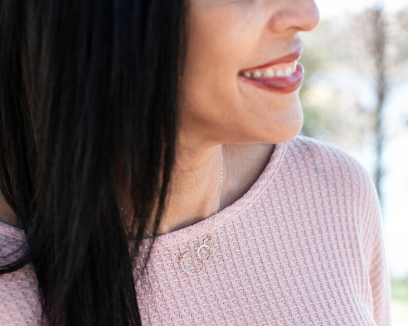 May include: A woman wearing a pink waffle knit sweater with a delicate gold necklace featuring two interlocking circles.
