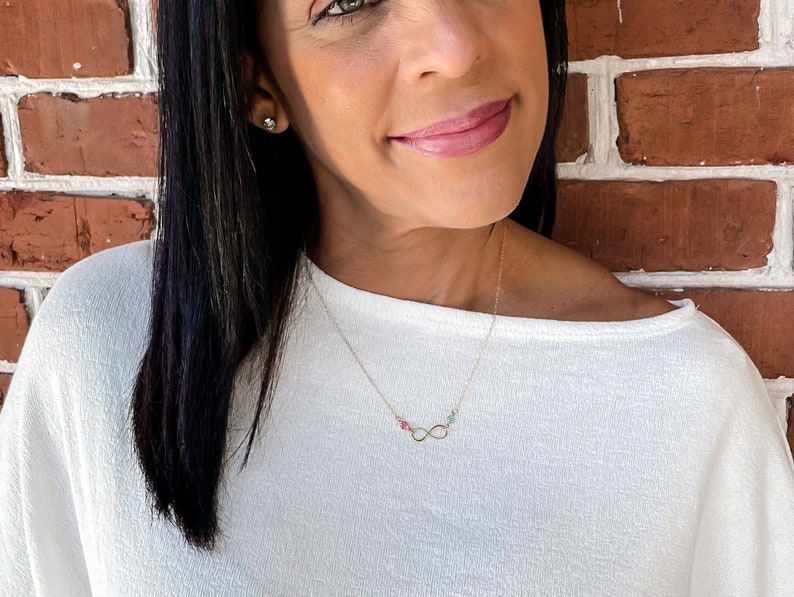 May include: A gold infinity necklace with small pink, green, and blue gemstones. The necklace is worn by a person with long black hair and a white shirt.