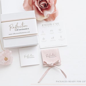 May include: A white gift box with the text "Reflection of memories" and a gold band. A gold necklace with a pendant is displayed next to a pink crystal. A small pink pouch and a card with jewelry care instructions are also visible.
