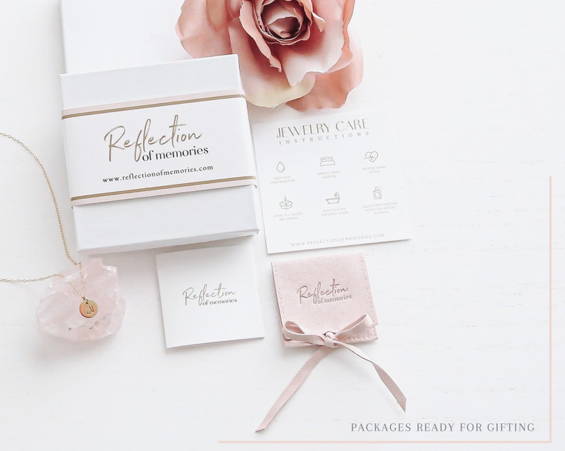 May include: A white gift box with the text "Reflection of memories" and a gold band. A gold necklace with a pendant is displayed next to a pink crystal. A small pink pouch and a card with jewelry care instructions are also visible.