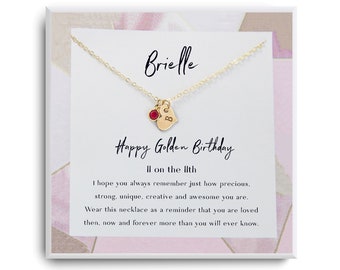 May include: A gold-colored necklace with a heart pendant and a small red gemstone. The necklace is on a white card with the text "Brielle" and "Happy Golden Birthday". The card has a pink and white geometric design.