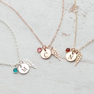 May include: Three delicate necklaces with angel wing charms and initial pendants. The necklaces are silver, rose gold, and gold. Each necklace features a small gemstone in a different color: turquoise, pink, and red.