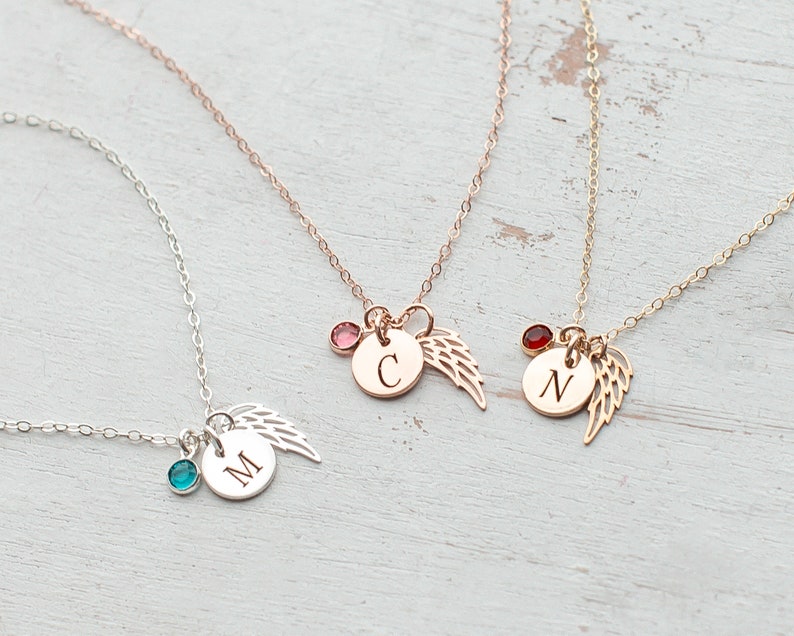 May include: Three delicate necklaces with angel wing charms and initial pendants. The necklaces are silver, rose gold, and gold. Each necklace features a small gemstone in a different color: turquoise, pink, and red.