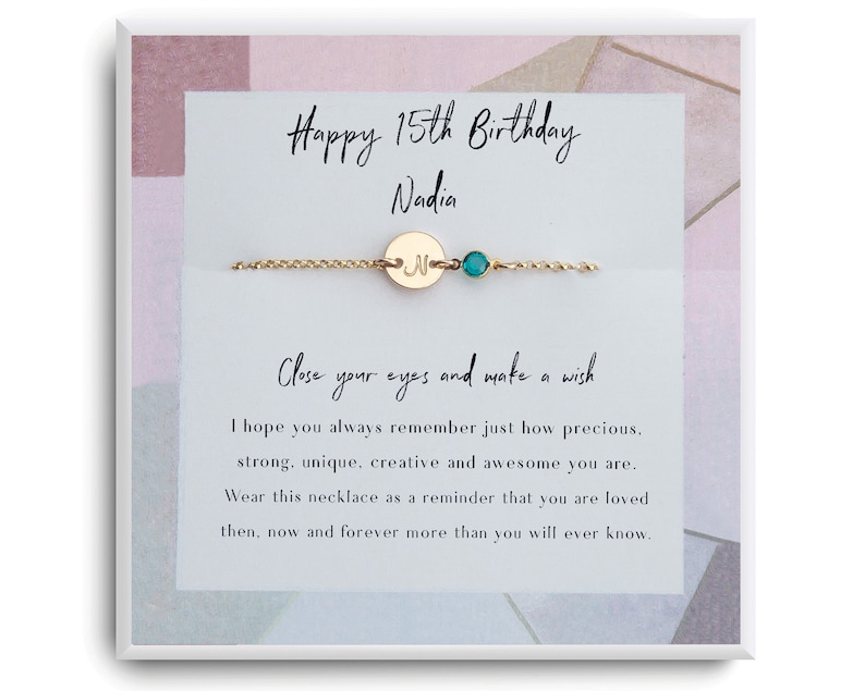 May include: A gold necklace with a small round charm that says "N" and a turquoise gemstone. The necklace is packaged in a white box with a pink and grey background. The box has the text "Happy 15th Birthday Nadia" and a message about making a wish.