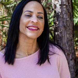 May include: A woman with long black hair is smiling and wearing a pink waffle knit top with a gold necklace. She is standing in front of a tree.