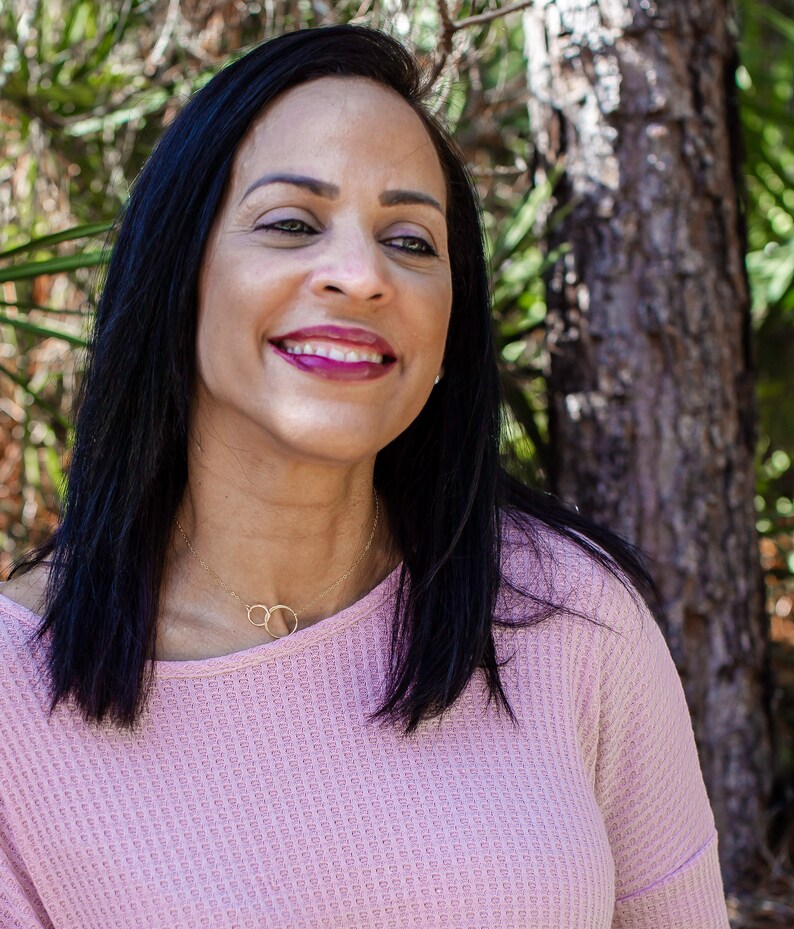 May include: A woman with long black hair is smiling and wearing a pink waffle knit top with a gold necklace. She is standing in front of a tree.