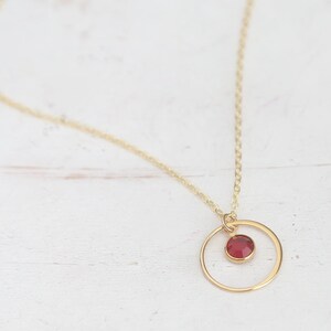 Puede incluir: Un collar de cadena dorada con un peque&ntilde;o colgante de piedra roja dentro de un c&iacute;rculo dorado.