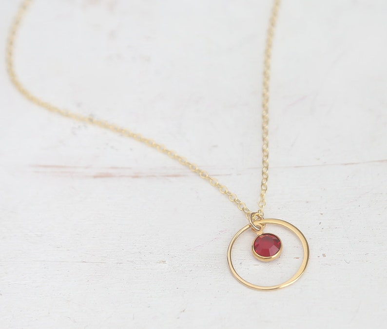 Puede incluir: Un collar de cadena dorada con un peque&ntilde;o colgante de piedra roja dentro de un c&iacute;rculo dorado.