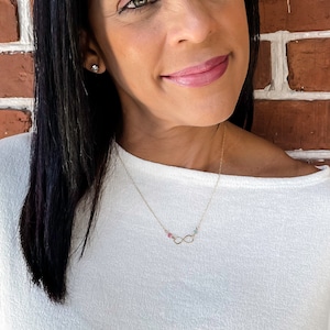 May include: A gold infinity necklace with small pink, green, and blue gemstones. The necklace is worn by a person with long black hair and a white shirt.