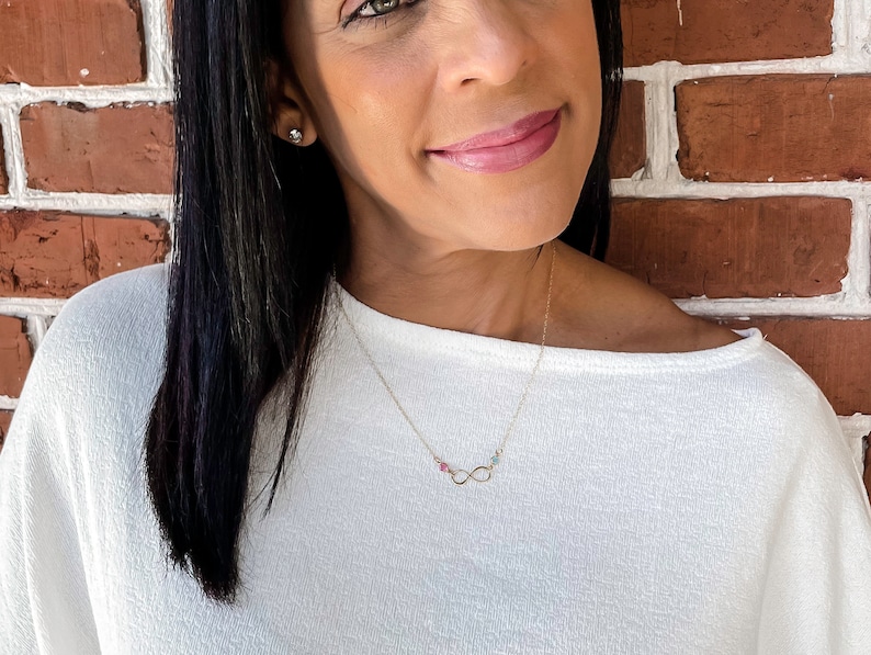 May include: A gold infinity necklace with small pink, green, and blue gemstones. The necklace is worn by a person with long black hair and a white shirt.