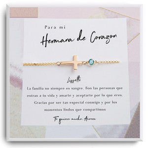 Könnte beinhalten: Ein goldfarbenes Kreuzarmband mit einem kleinen blauen Edelstein. Das Armband wird auf einer weißen Karte mit spanischem Text präsentiert, einschließlich "Para mi Hermana de Corazon" und einer persönlichen Nachricht.