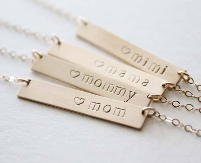 May include: Four gold bar necklaces with the words "mom", "mommy", "mama", and "mini" engraved on them. Each bar has a small heart symbol before the word.