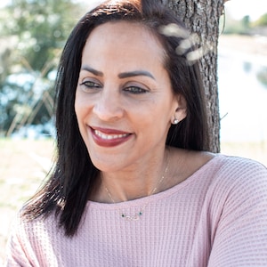 May include: A woman with long dark hair is wearing a pink sweater and a gold necklace with a small infinity symbol and green stones. She is smiling and looking to the side.