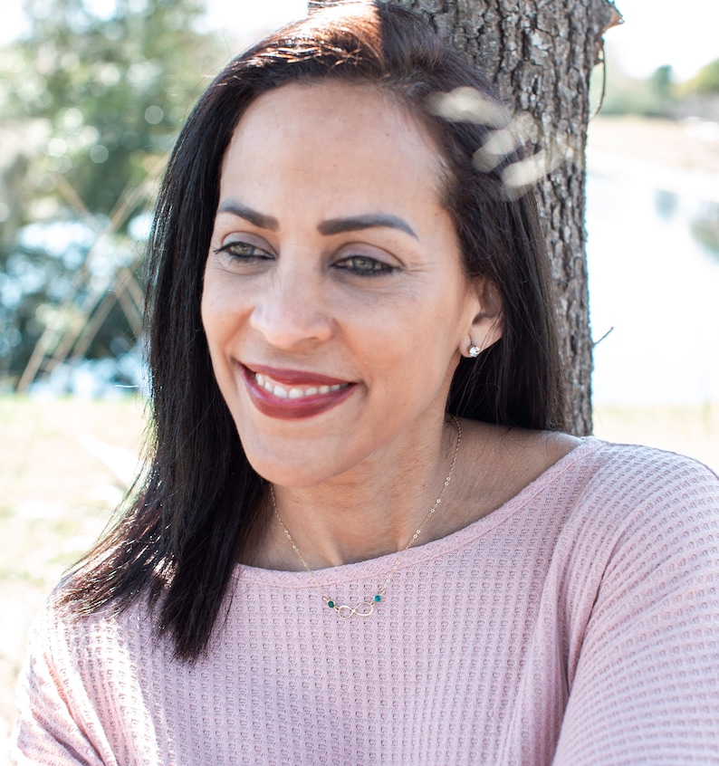 May include: A woman with long dark hair is wearing a pink sweater and a gold necklace with a small infinity symbol and green stones. She is smiling and looking to the side.