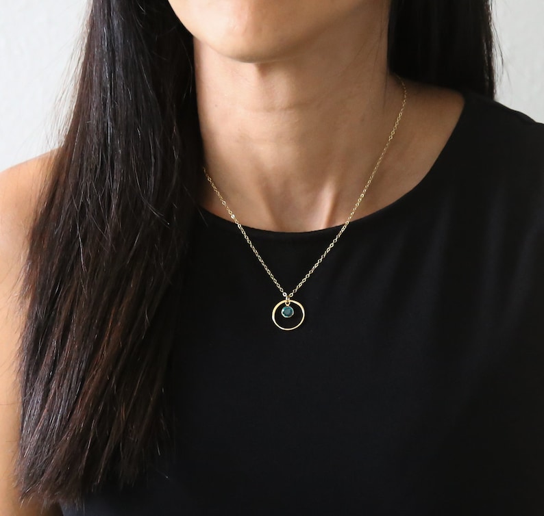 Puede incluir: Un collar de cadena dorada con un peque&ntilde;o colgante redondo de piedra preciosa azul verdoso que cuelga de un fino c&iacute;rculo dorado.