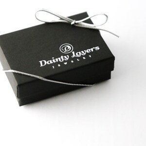 May include: A black jewelry box with a white ribbon and the text "Dainty Layers Jewelry" printed on the top.