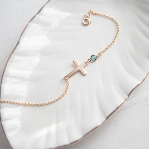 Puede incluir: Una pulsera dorada con un colgante de cruz y una peque&ntilde;a gema azul redonda. La pulsera se muestra sobre un plato blanco con borde festoneado. La cruz est&aacute; en posici&oacute;n horizontal y la gema est&aacute; cerca del cierre.
