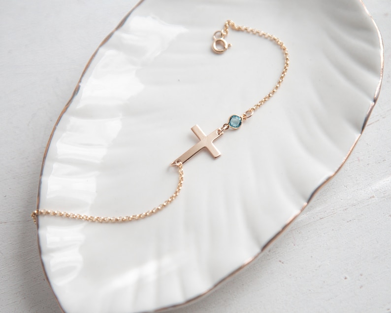 Puede incluir: Una pulsera dorada con un colgante de cruz y una peque&ntilde;a gema azul redonda. La pulsera se muestra sobre un plato blanco con borde festoneado. La cruz est&aacute; en posici&oacute;n horizontal y la gema est&aacute; cerca del cierre.